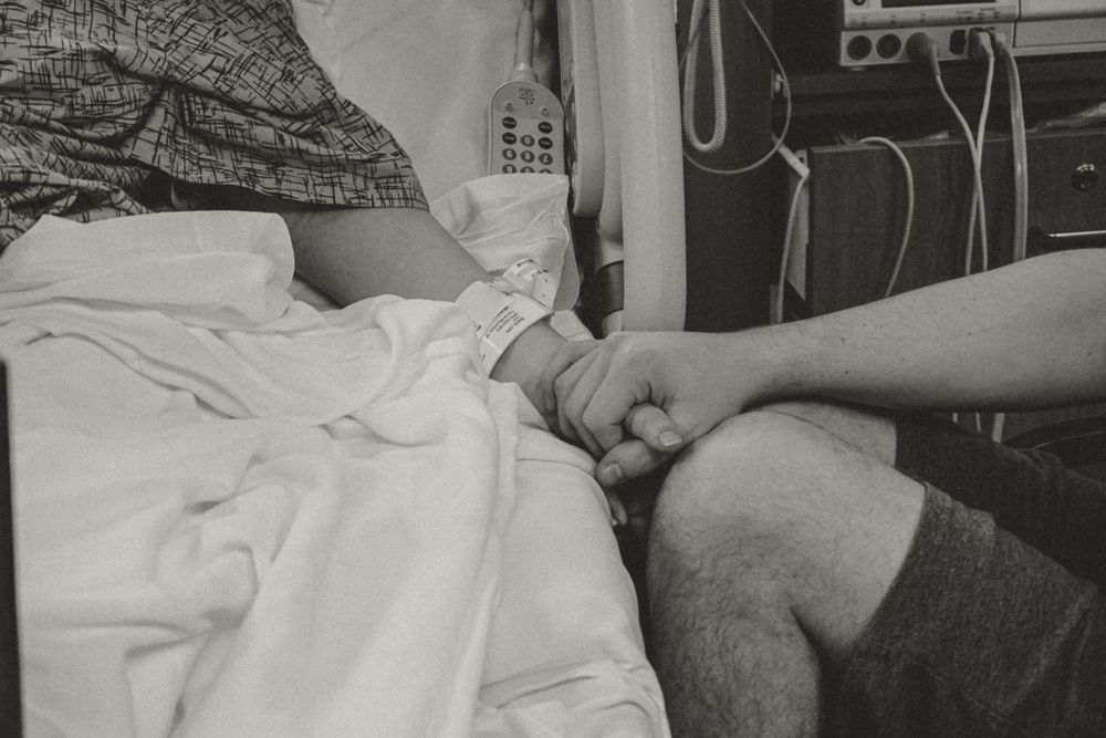 Black and white hospital photograph showing a patient resting in a bed with medical equipment visible in the background.