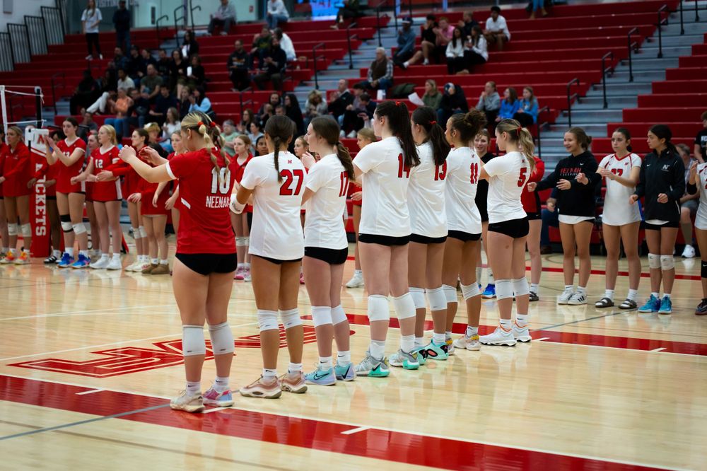 Volleyball team lines up on court during pre-game ceremony in gymnasium with red bleachers.