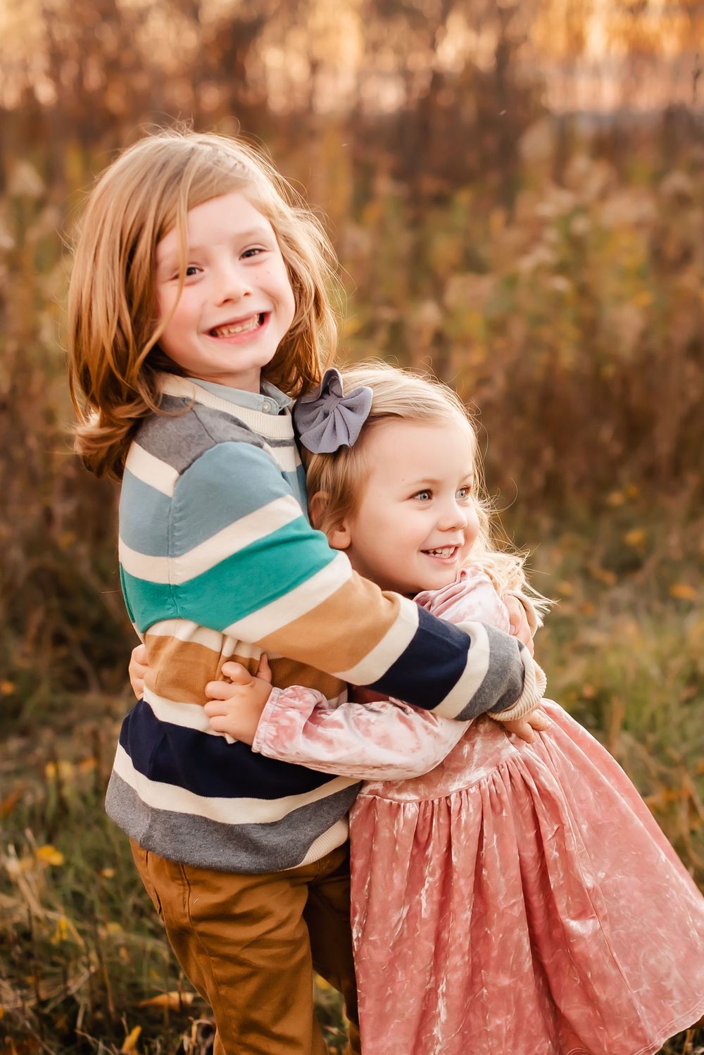 Two young children hug and laugh together outdoors in autumn wearing coordinating outfits.