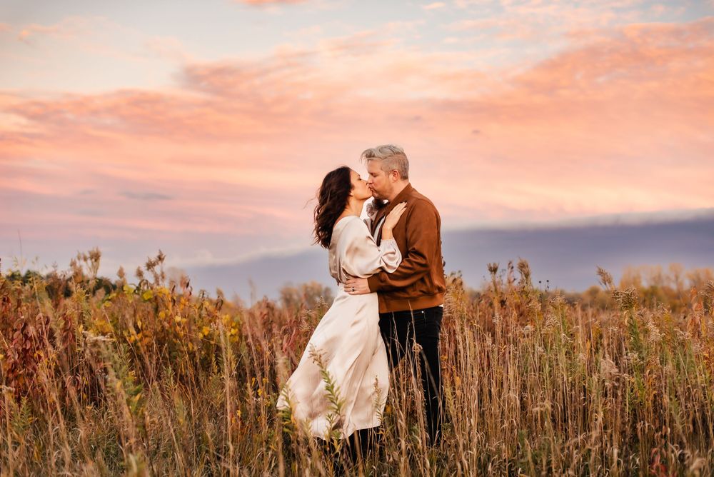 A couple embraces in a golden field of tall grass during sunset with pink and orange sky in the background.