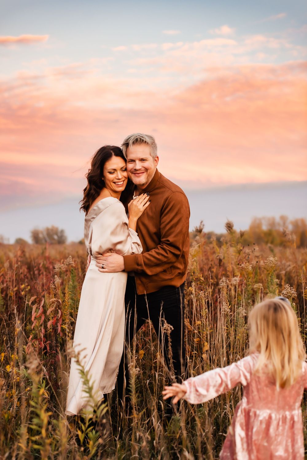 A couple embraces in a field of tall grass and wildflowers at sunset while a child in pink reaches towards them.