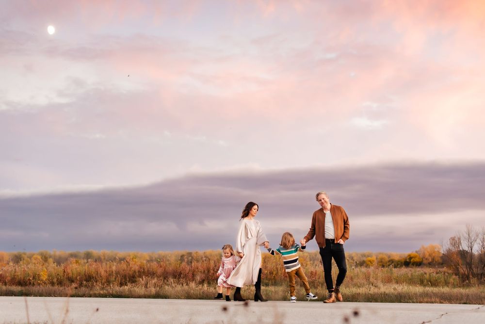 A family walks together on a path during golden hour with autumn foliage and pink clouds in the background.