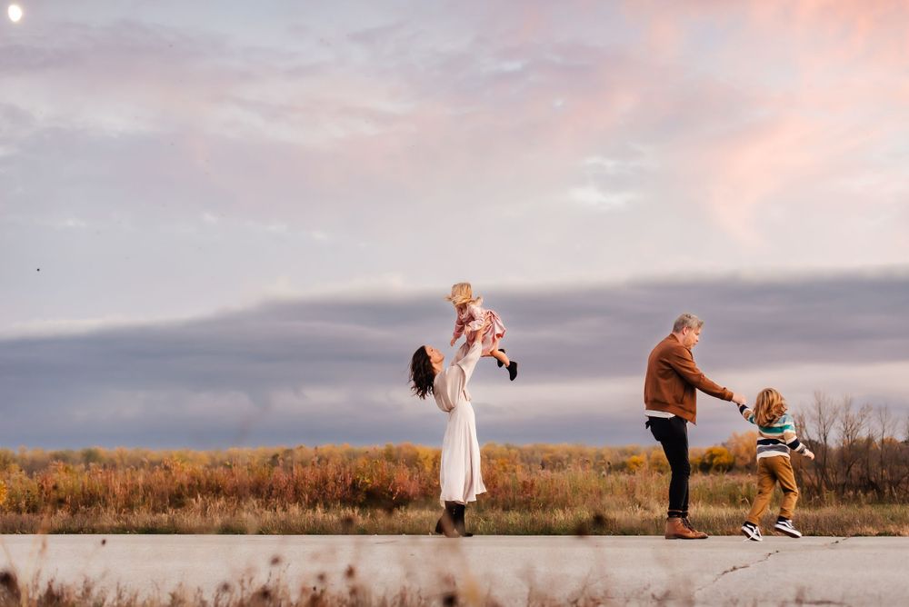 A couple lifts their children playfully against a scenic sunset backdrop with golden grasses and cloudy skies.