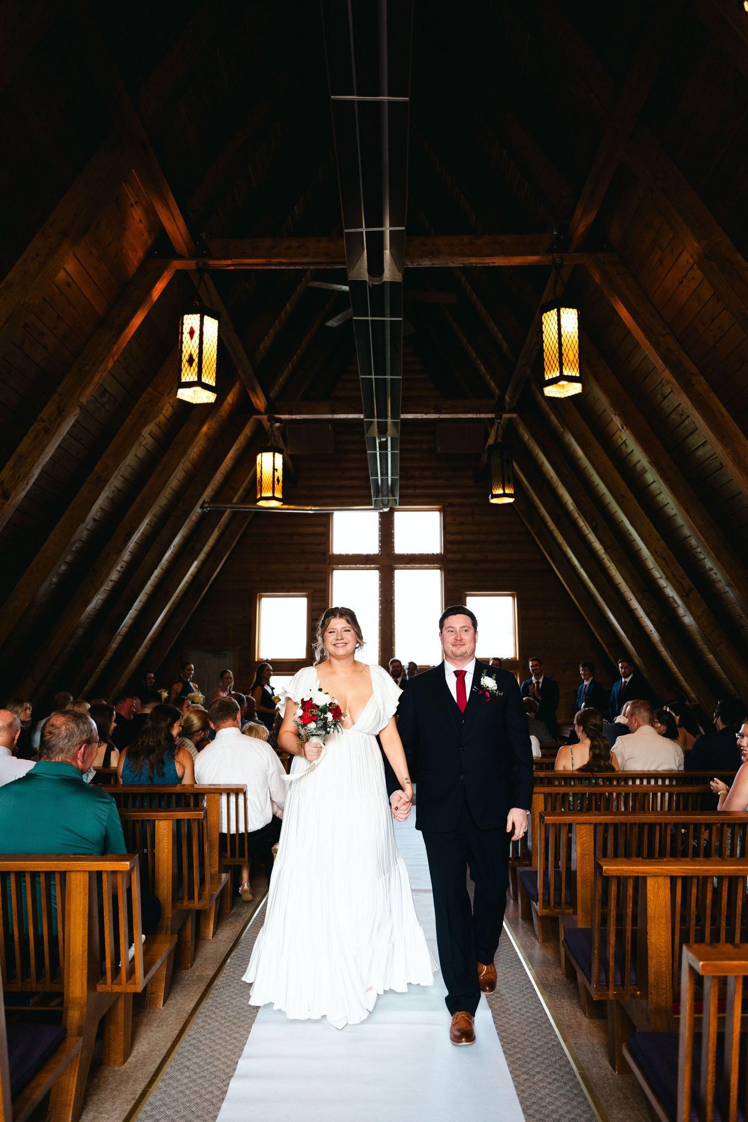 A couple walks down the aisle in a rustic wooden chapel with glowing lanterns and wooden pews.