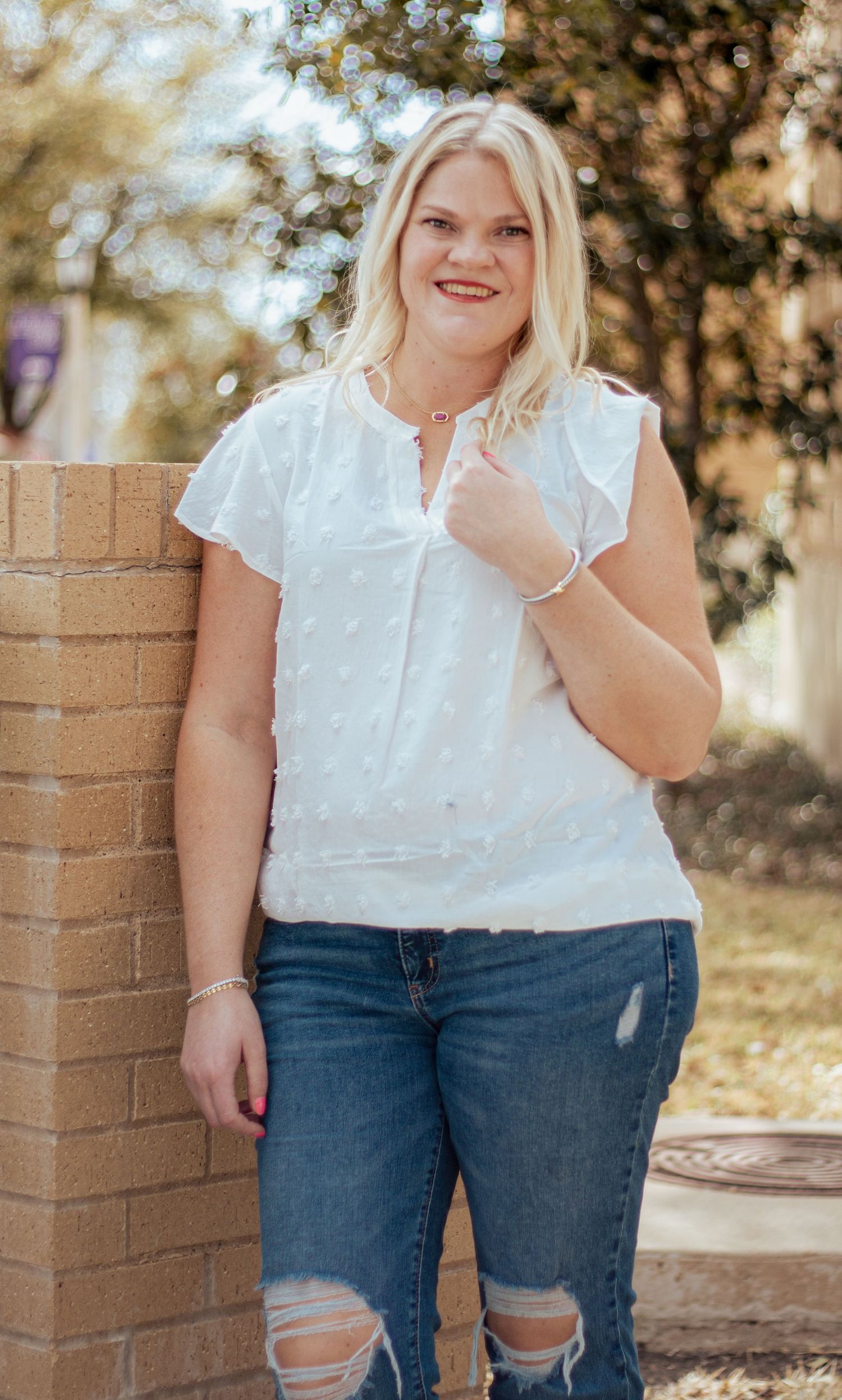 A person wearing a white blouse and distressed jeans leans against a brick wall outdoors on a sunny day.