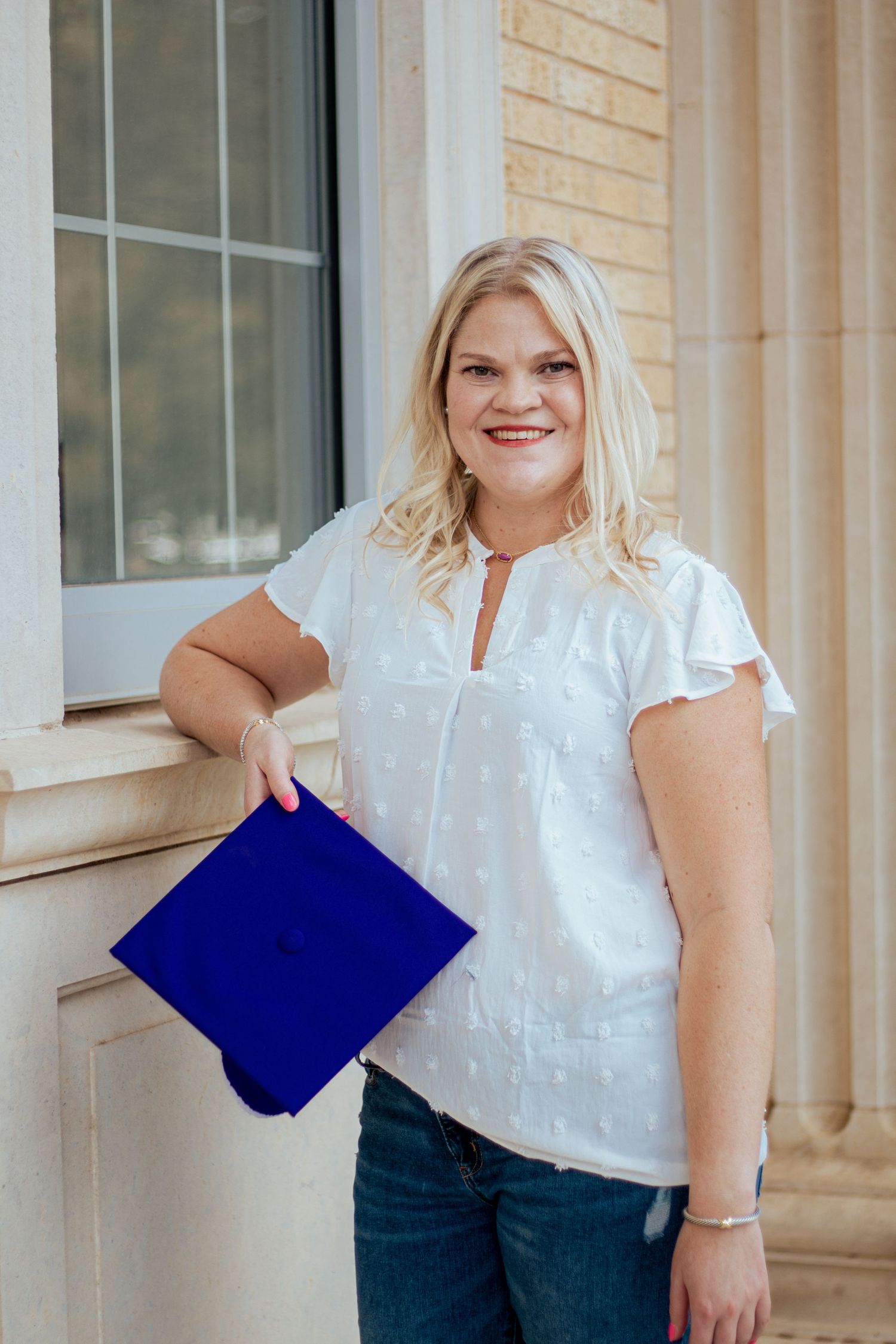 Graduate holding blue diploma folder stands smiling in white blouse and jeans against brick building exterior.