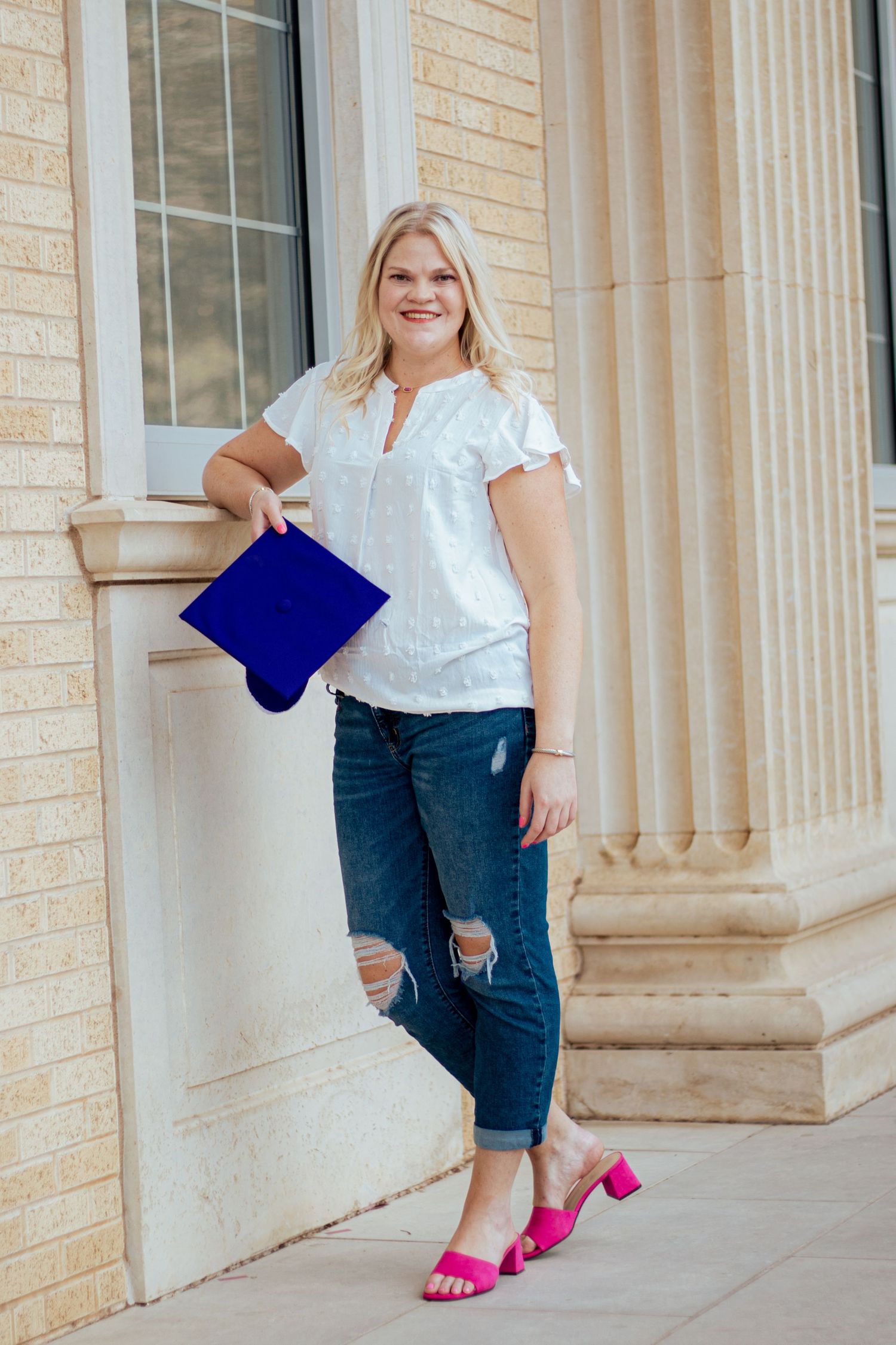 A woman in a white top stands against a stone column wearing ripped jeans and bright pink sandals while holding a blue clutch.
