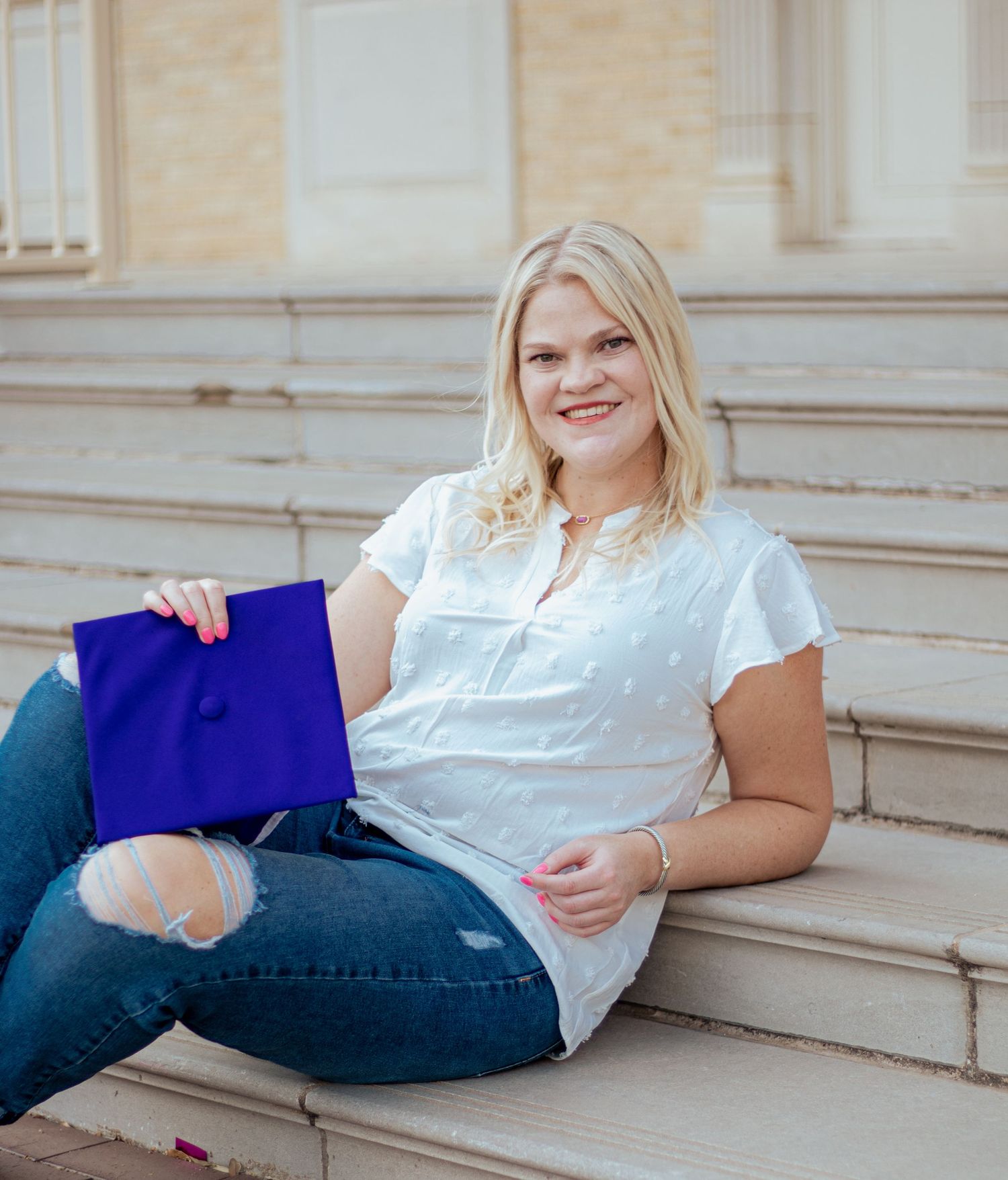 A person in ripped jeans and white shirt sits casually on stone steps holding a bright blue shopping bag.