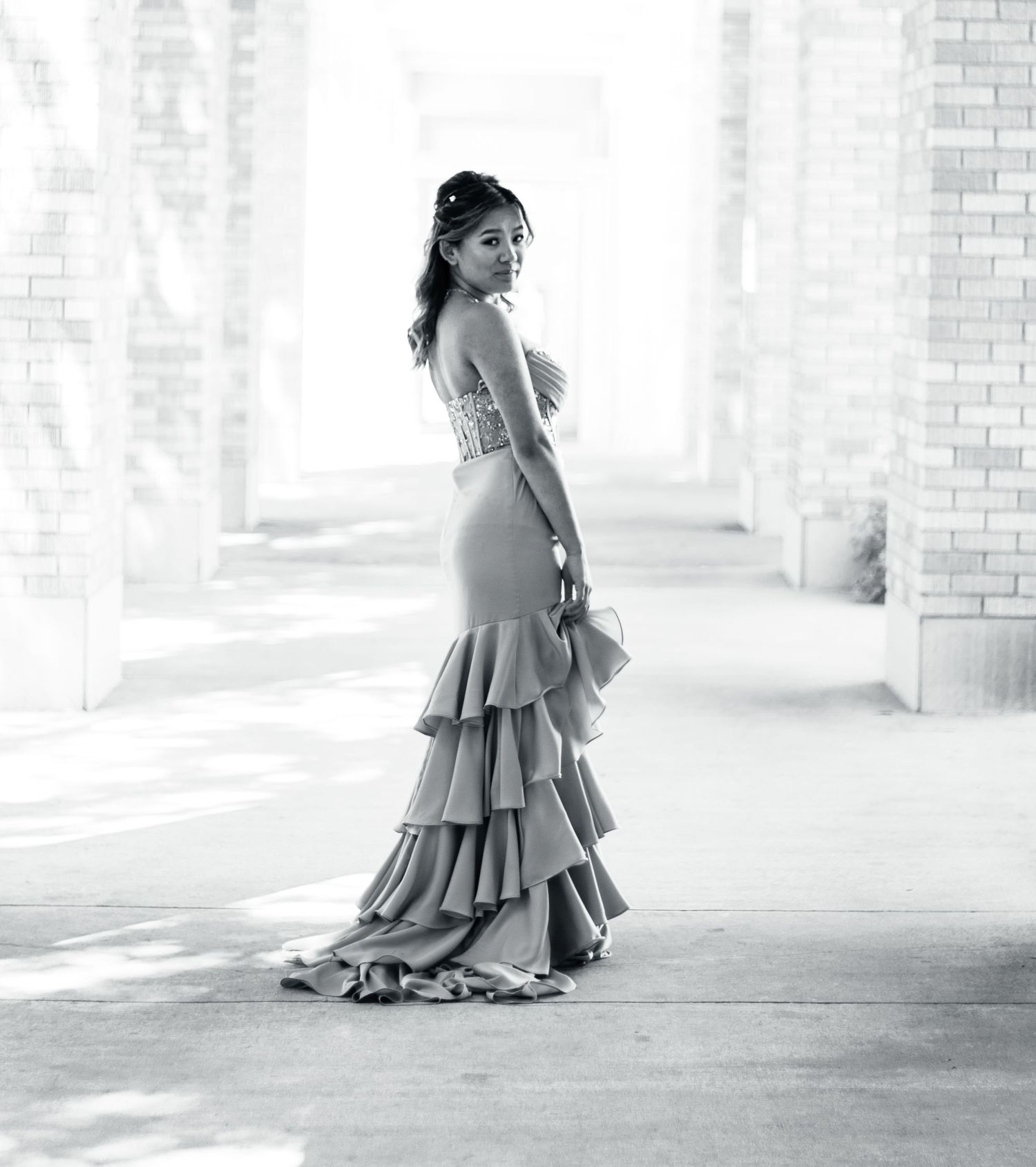 A flowing ruffled evening gown with tiered skirt photographed in dramatic black and white lighting in a brick corridor.