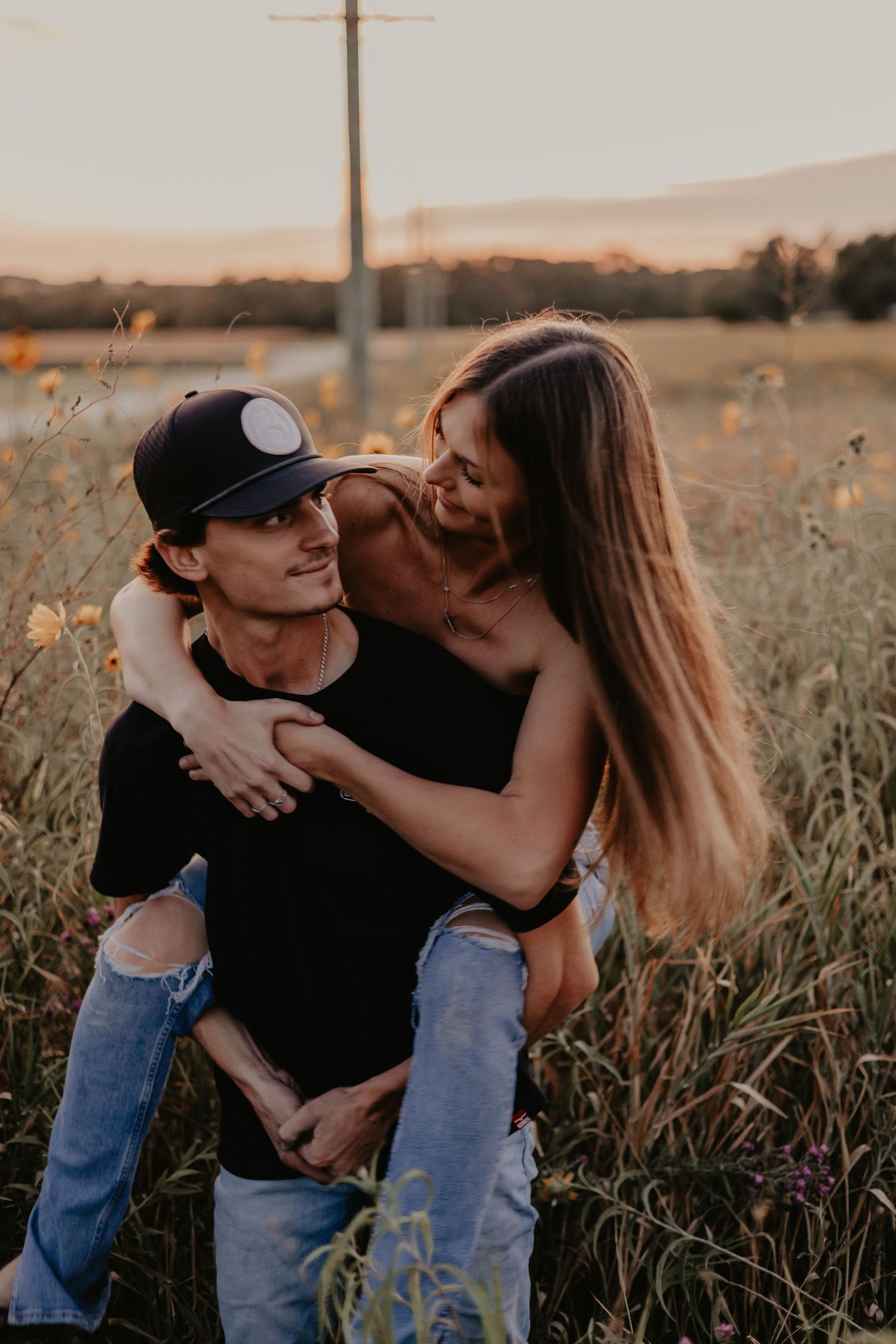 A couple shares a tender moment at sunset in a wildflower field, with one giving the other a piggyback ride.