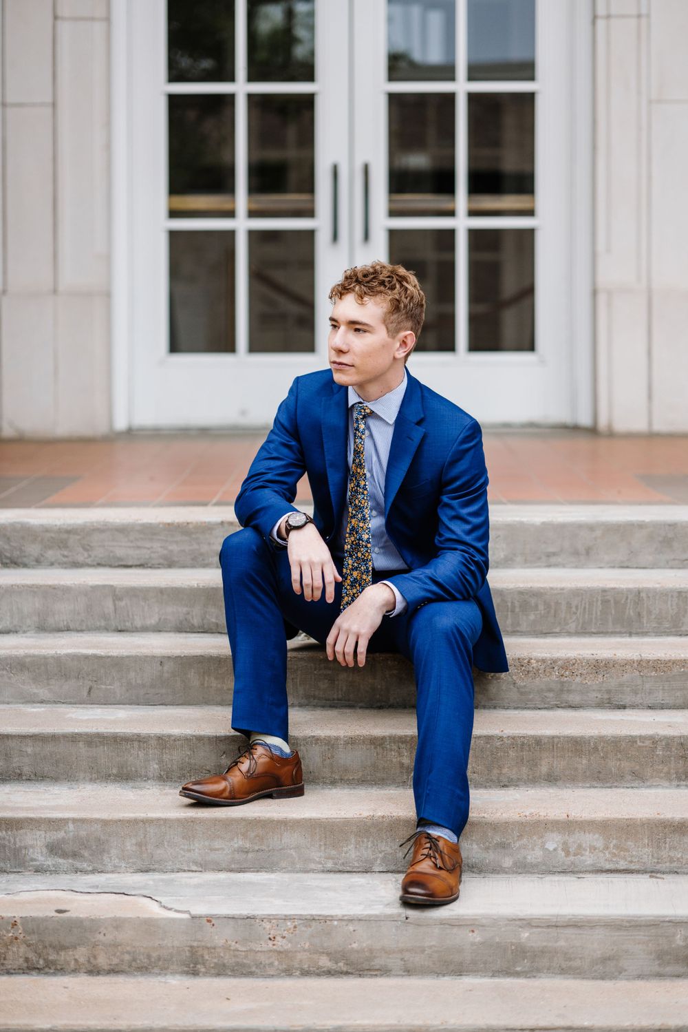 Young person in a navy blue suit sits casually on concrete steps wearing brown dress shoes.