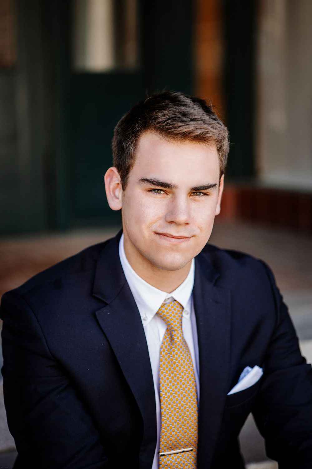 Person in navy suit with yellow tie poses formally against dark background.