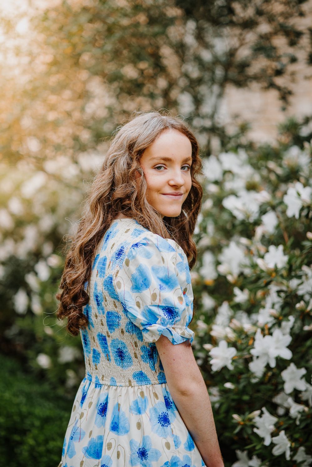 Person in blue floral dress poses gracefully against backdrop of white flowers and greenery.