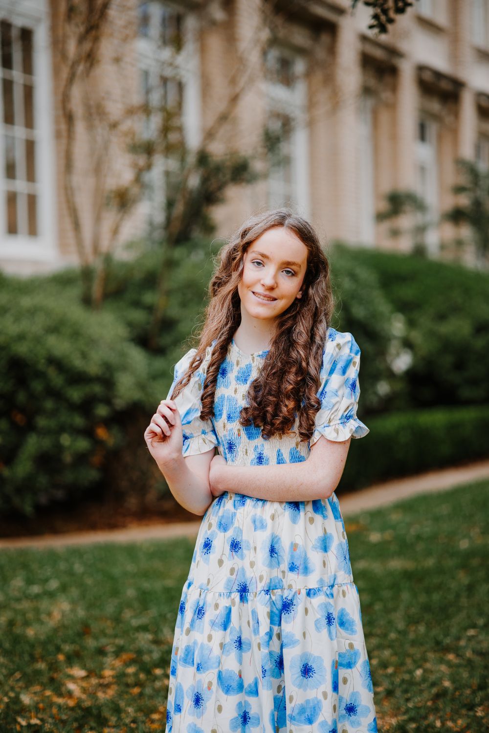 Person in blue and white floral dress poses playfully on grassy lawn near building.
