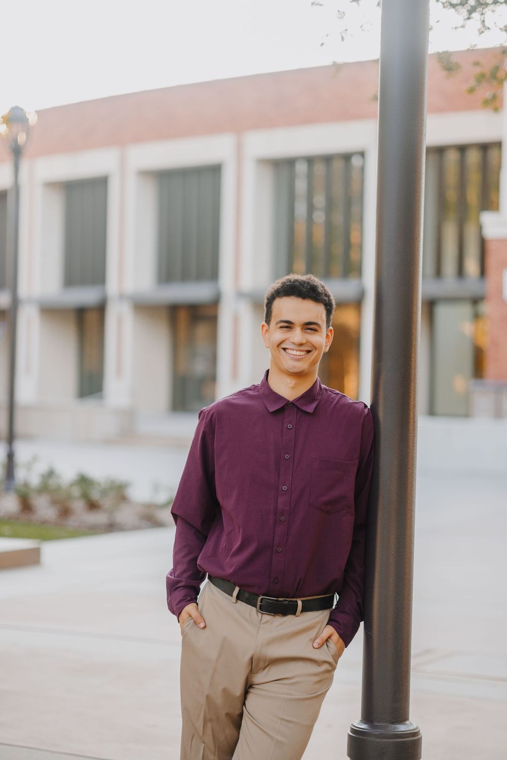 A person wearing a burgundy polo shirt and khaki pants stands casually outdoors near architectural columns.