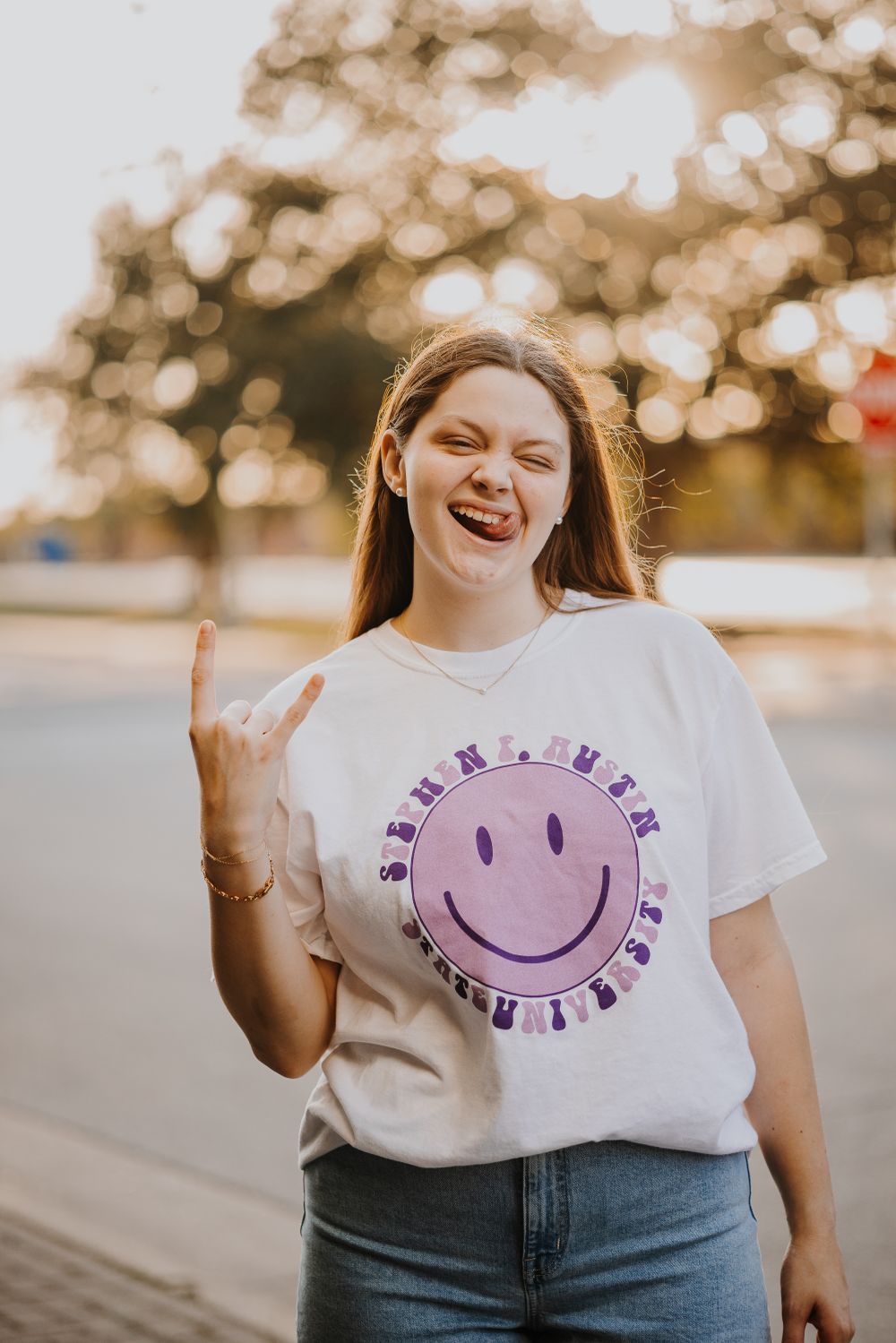 Person wearing white t-shirt with purple smiley face design smiles joyfully outdoors.