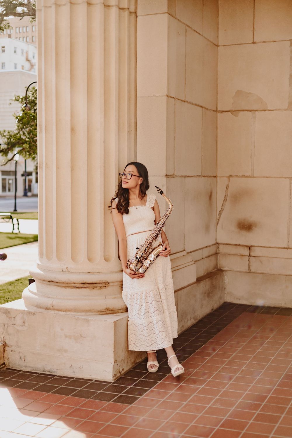 A person in a white dress poses playfully between elegant stone columns on a terracotta tiled porch.
