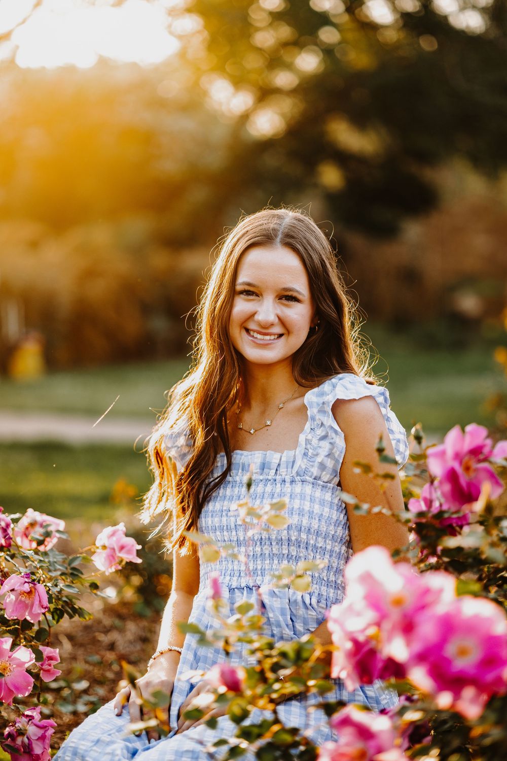 A person in a blue and white gingham dress poses joyfully among vibrant pink flowers during golden hour sunset.