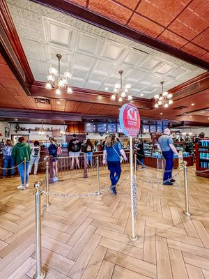 Customers wait in line at a restaurant counter with herringbone floors and vintage-style chandeliers overhead.