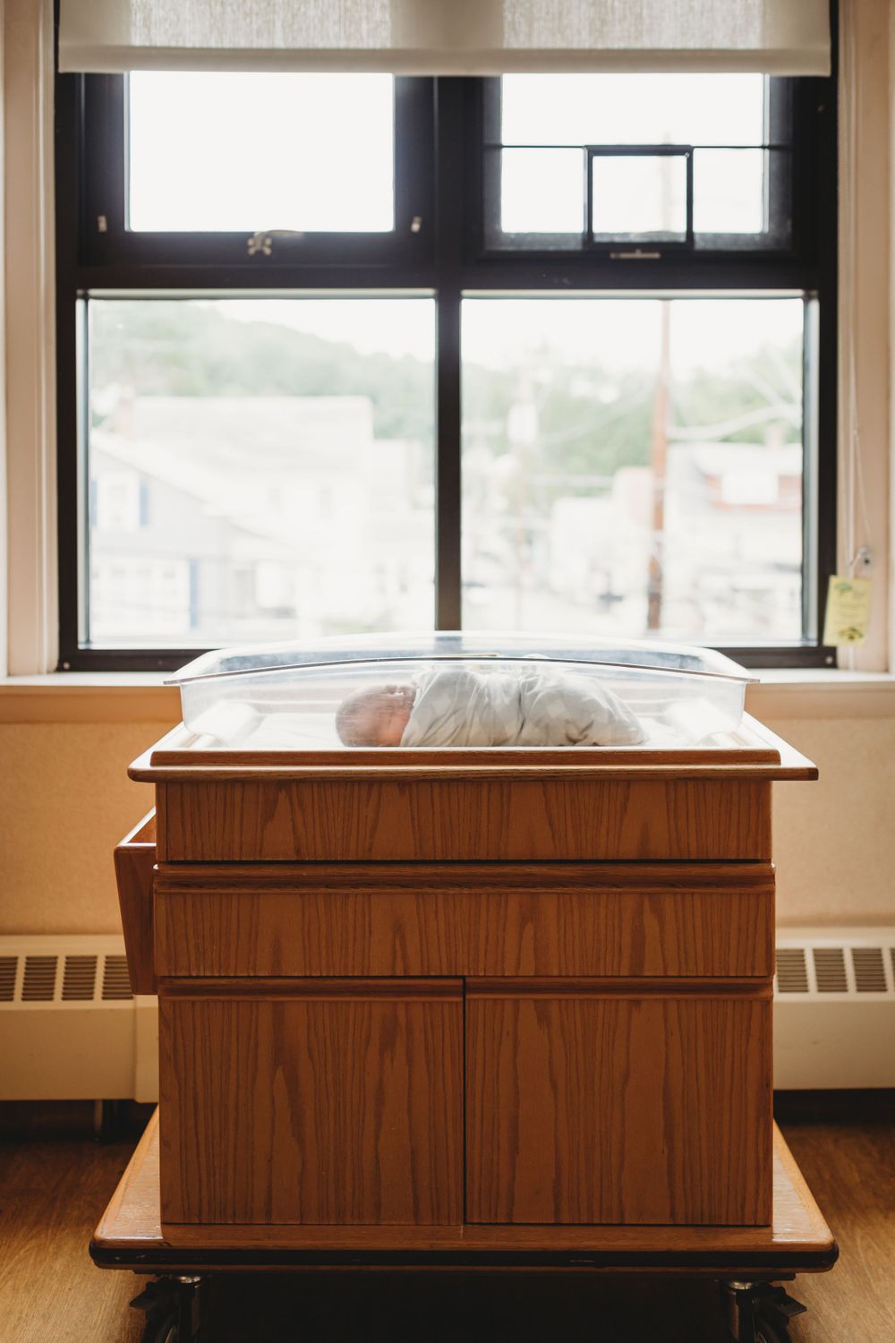 Wooden hospital bassinet near window with swaddled newborn.