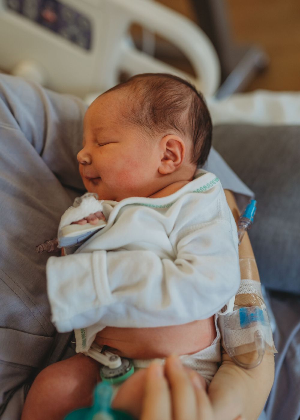 Newborn sleeping peacefully in white onesie on grey couch with medical equipment.