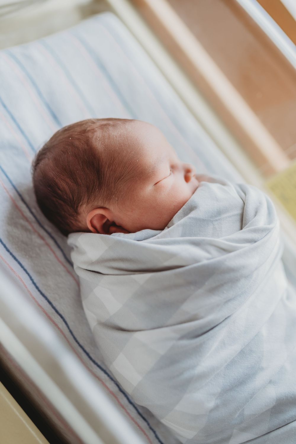 Peaceful newborn wrapped in soft grey swaddle sleeping in hospital bassinet.