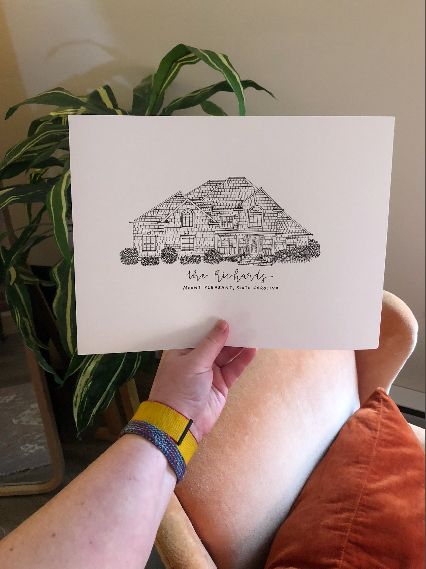 Hand holding a detailed architectural pen sketch of a two-story suburban house with landscaping details.