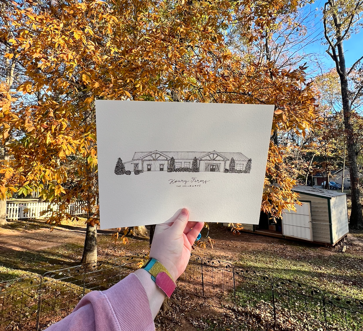 Hand holding architectural sketch of a single-story ranch house against autumn trees.