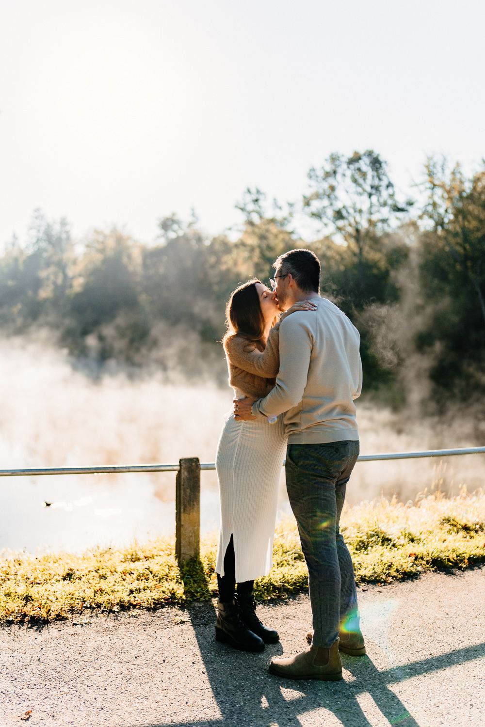 Couple sharing an intimate moment by a misty lake at sunrise while leaning against a wooden fence.