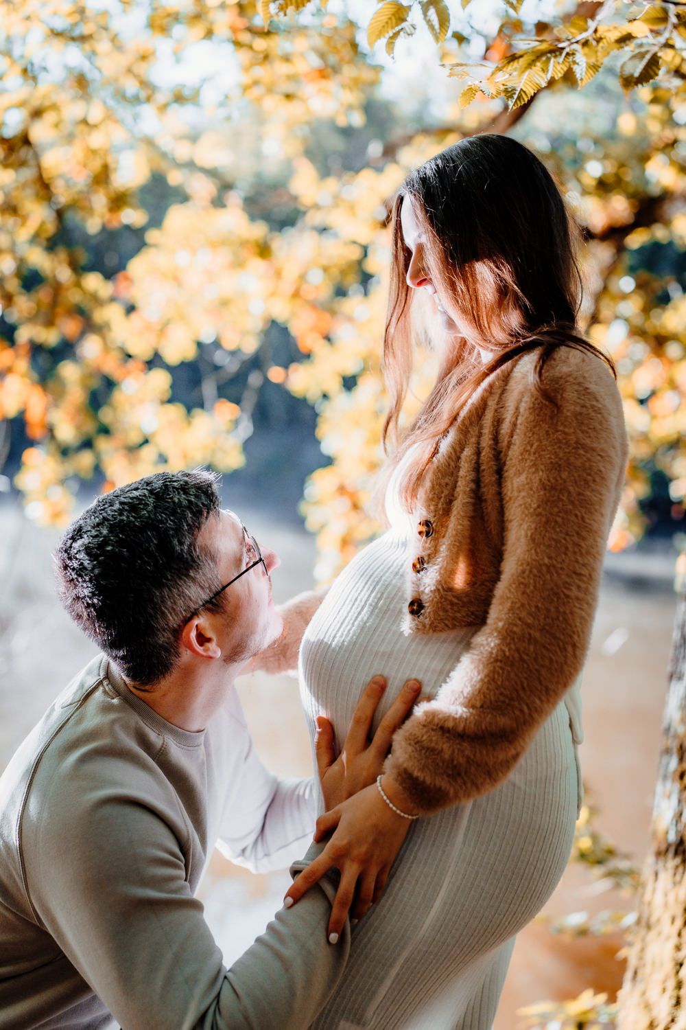 A tender maternity photoshoot moment outdoors with fall foliage in the background.