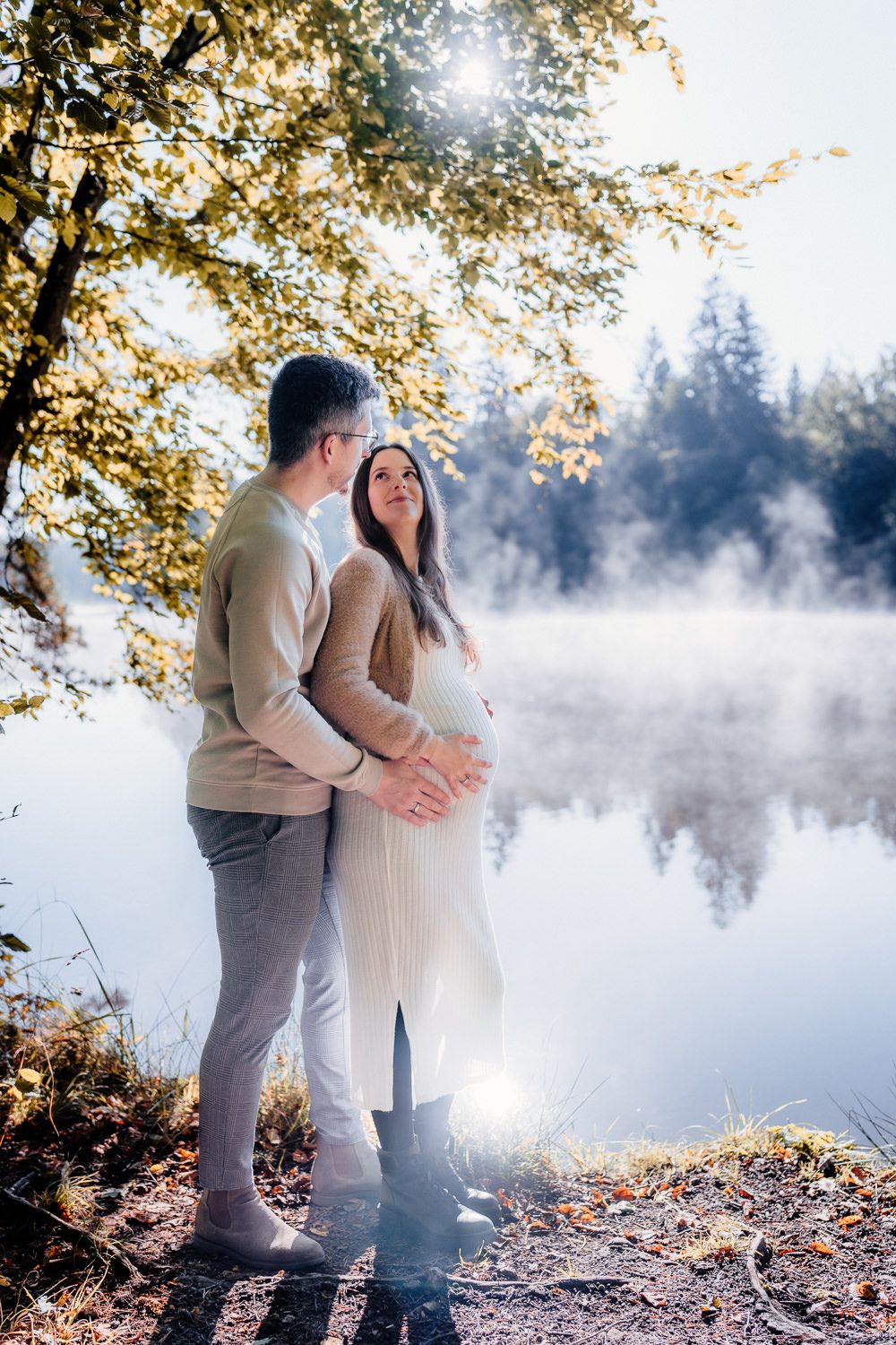 A romantic maternity photoshoot by a foggy lake with fall foliage in the background.