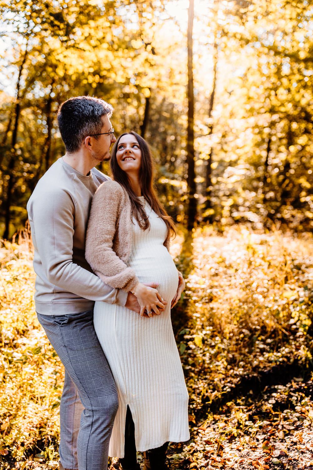A maternity photo showing a couple embracing in golden forest light.