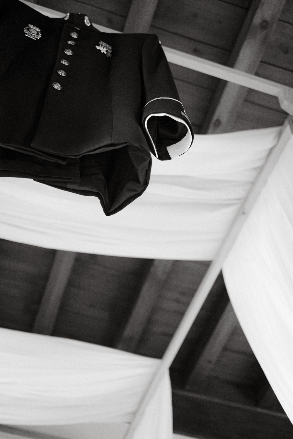 Black military jacket hanging from ceiling with white fabric draping in background.