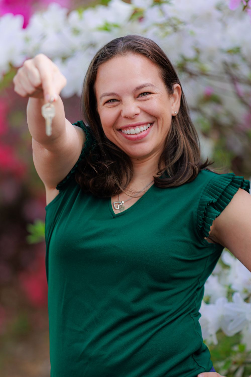 Smiling woman in green top holds out a key toward camera with pink flowers in background.