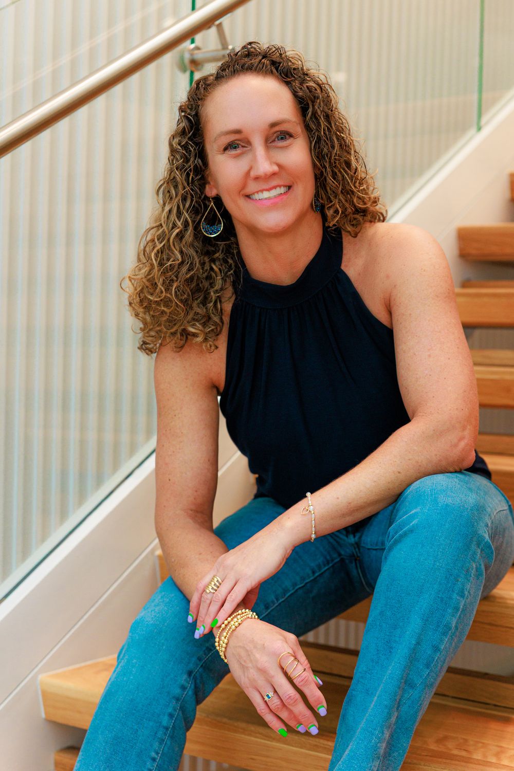 Smiling woman with curly hair sits on wooden stairs wearing black halter top and jeans with gold jewelry.