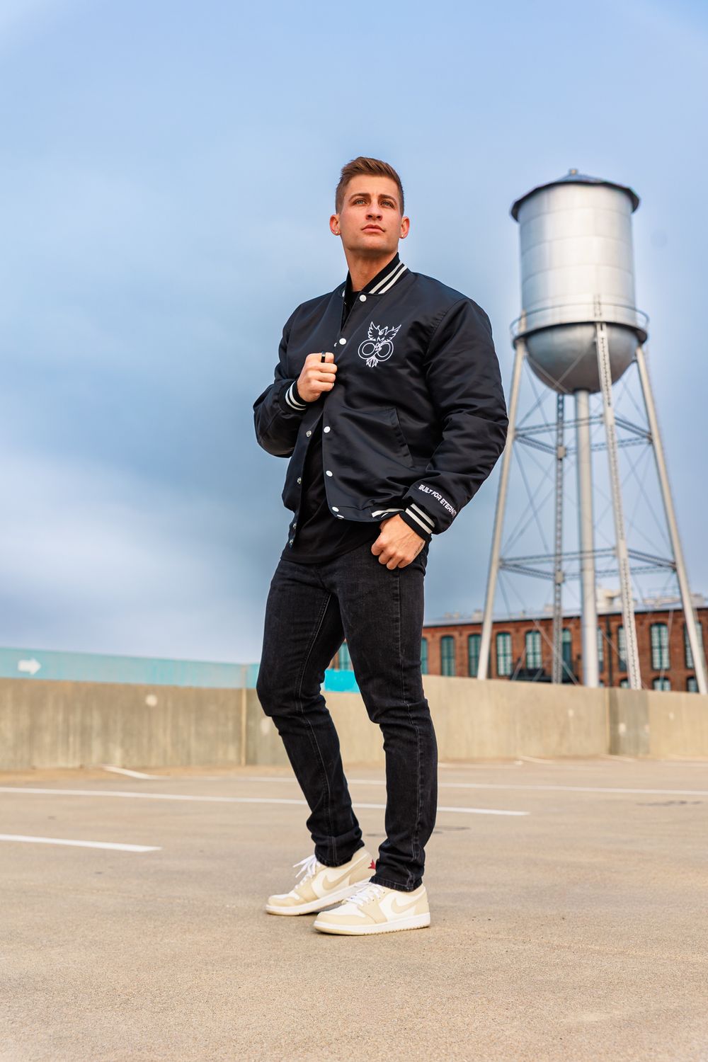 Person in black varsity jacket and pants poses against industrial backdrop with water tower in multiple angles.