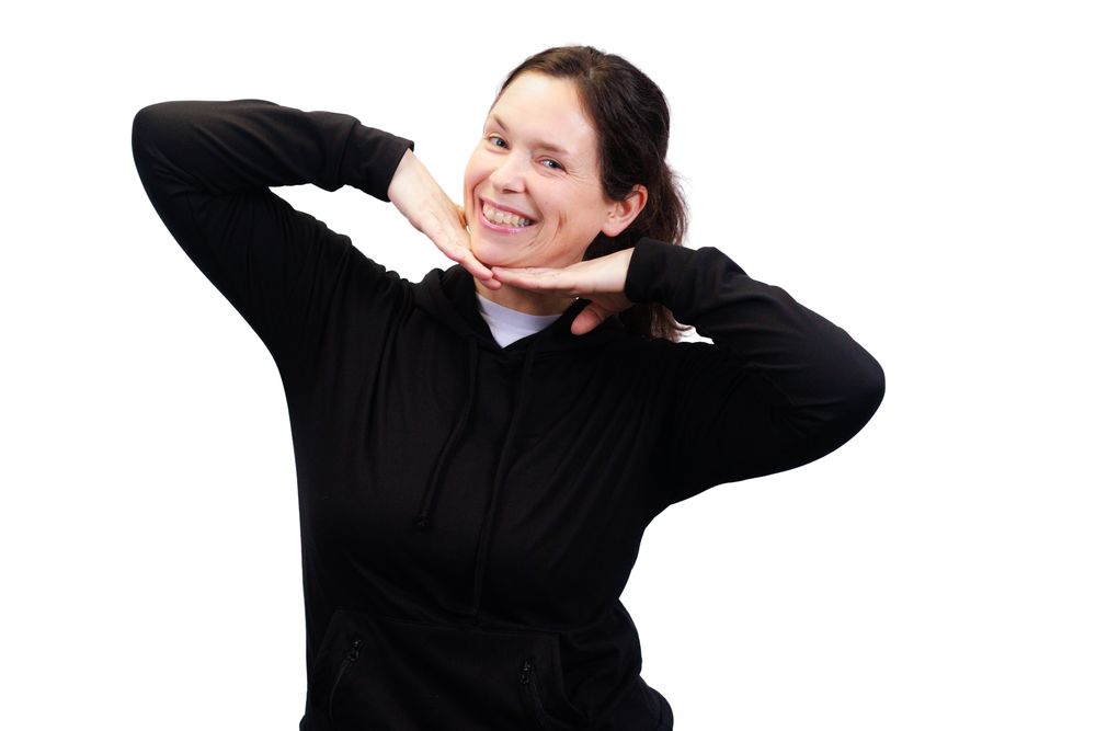 A person wearing a black sweater smiles and poses with hands behind their head against a white background.