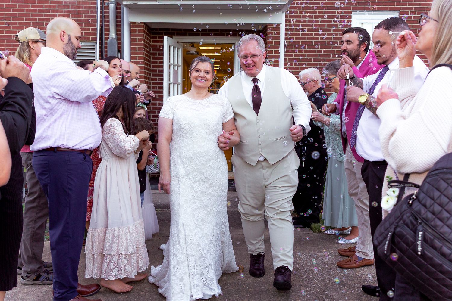 A couple exits a brick building while guests blow bubbles during a wedding celebration.