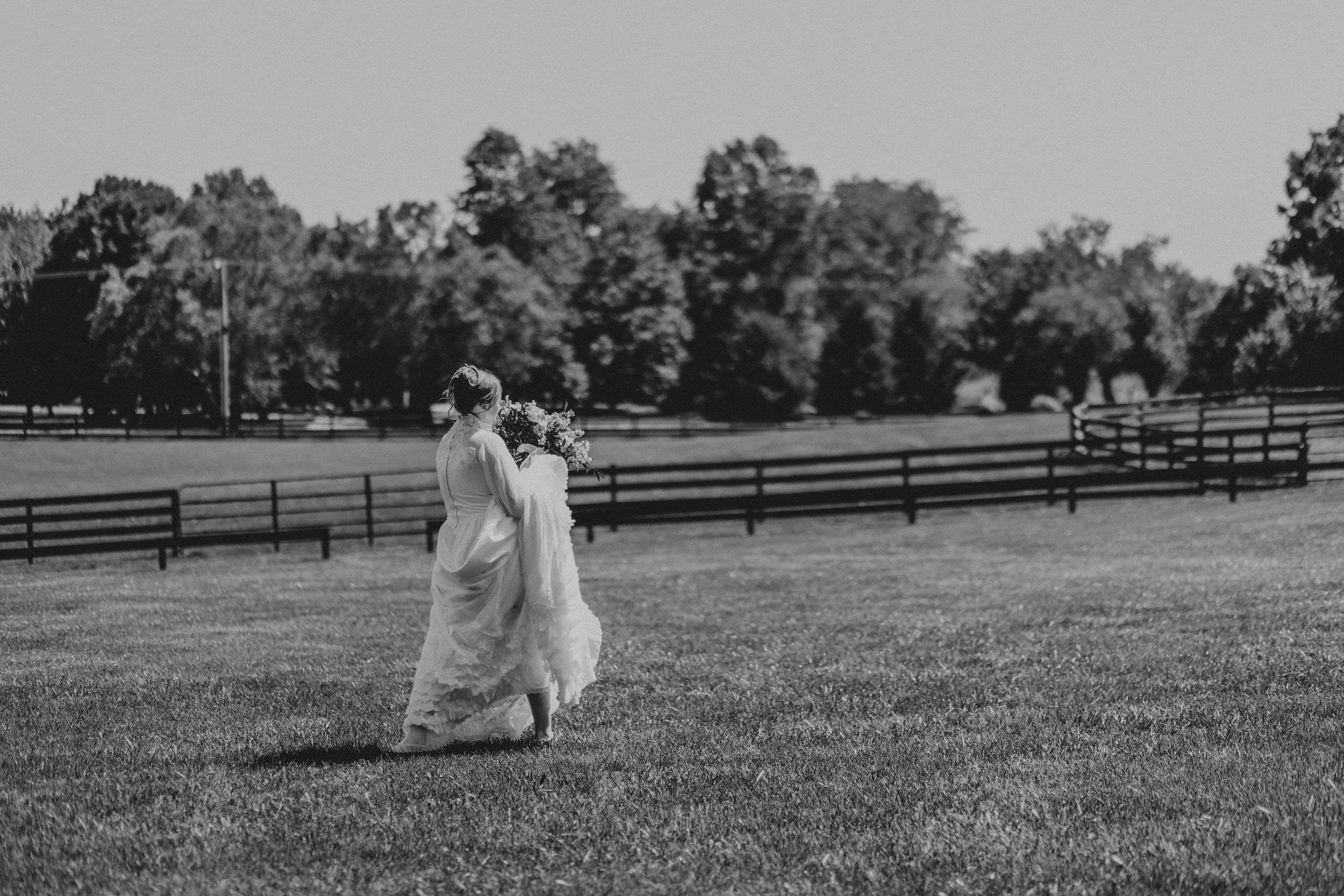 Two figures in flowing dresses walk across a grassy field bordered by wooden fences and trees in a black and white scene.