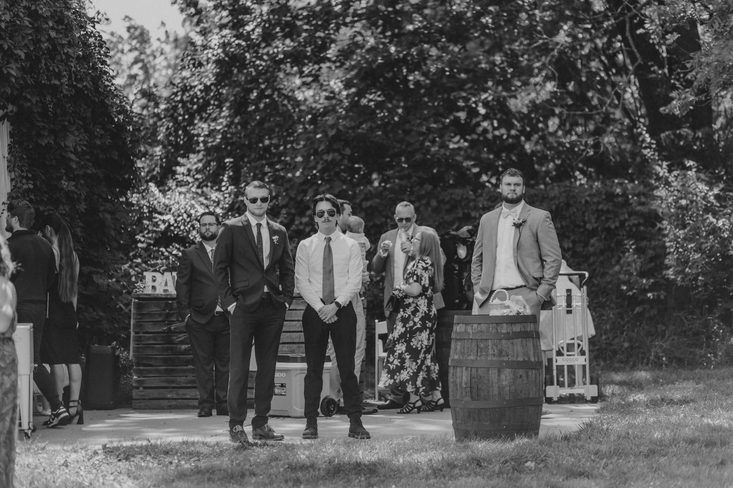 A vintage black and white photograph of a group of people standing outdoors near trees and a wooden barrel.