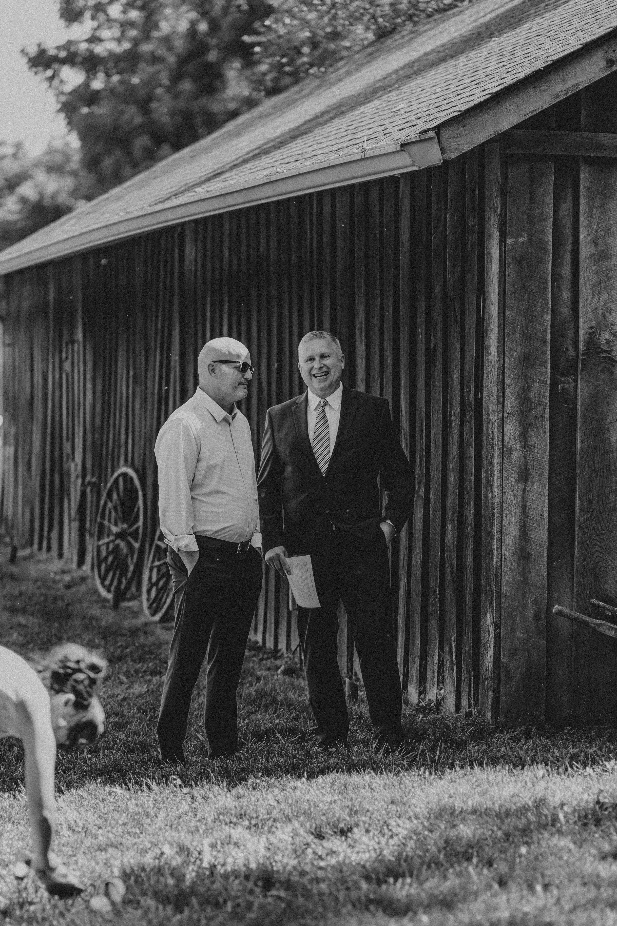 Two men stand in front of a rustic wooden barn, with a wagon wheel visible nearby and a dog in the foreground.