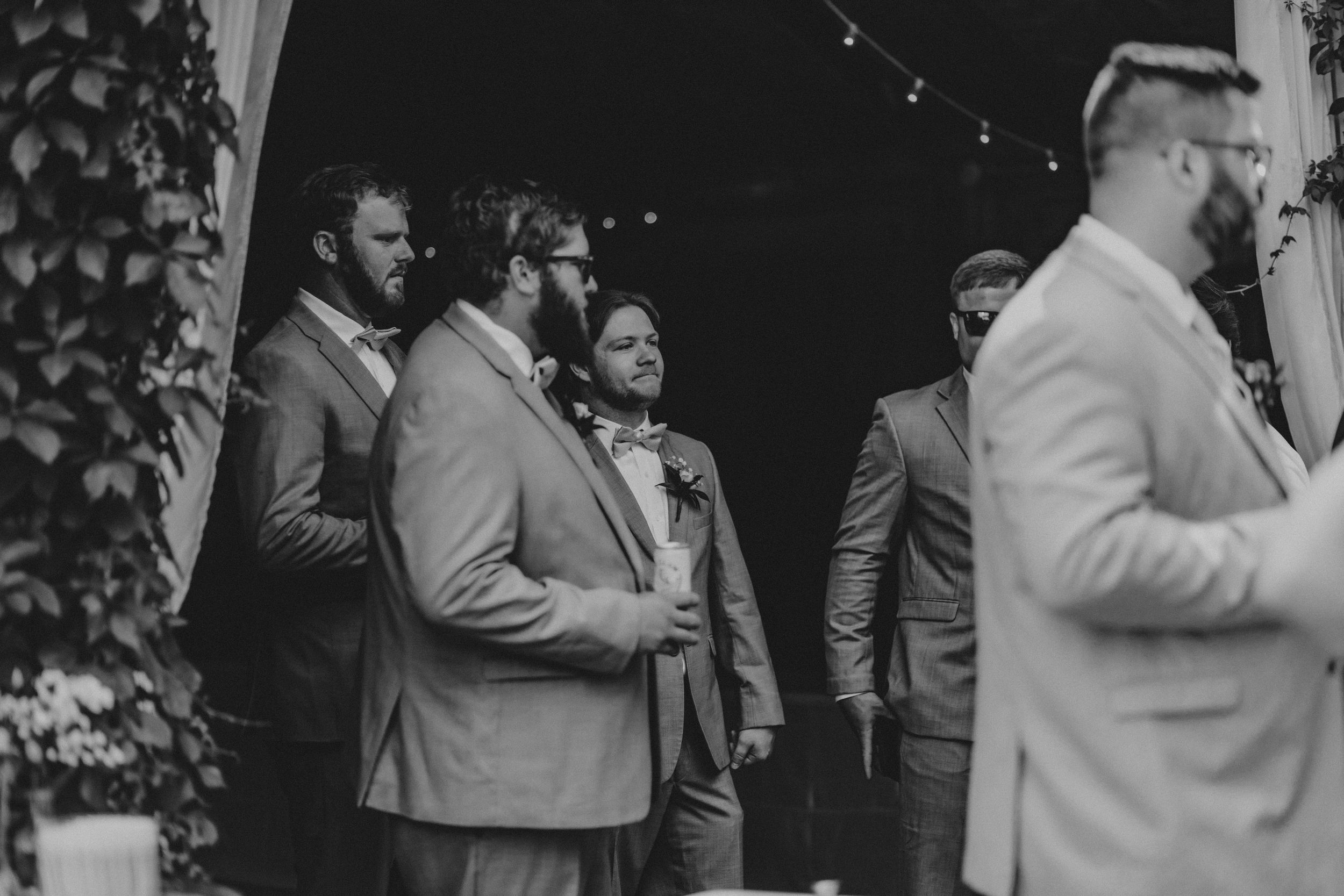Black and white photo of a group of men in suits standing together at an event with string lights visible.