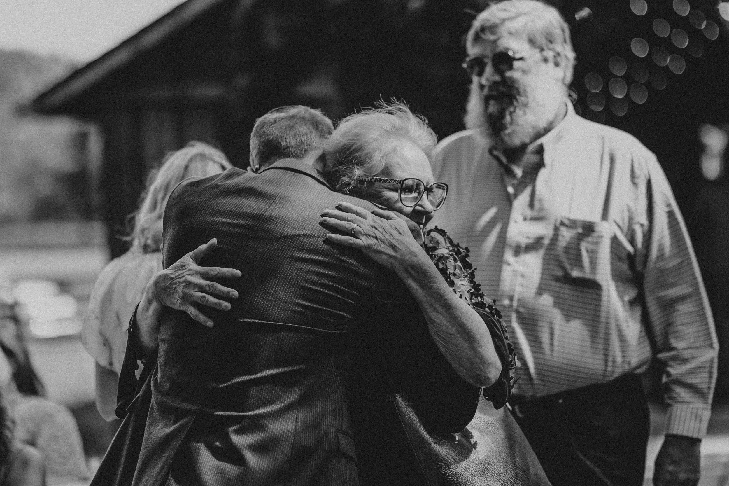 A black and white photograph captures an emotional embrace between people outdoors during a daytime gathering.