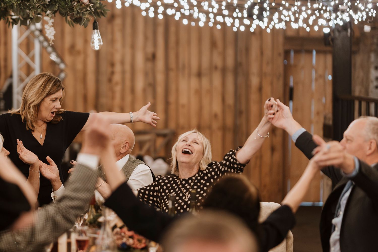 Wedding guests singing and celebrating under string lights at an indoor reception venue with wooden walls at Stanford Farm