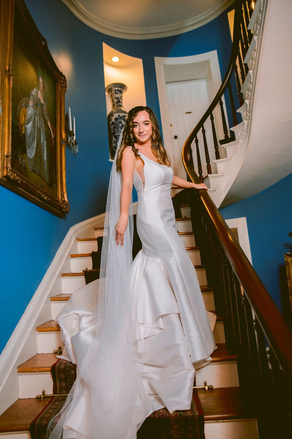 Wedding dress displayed on wooden staircase with dark banister against blue walls.