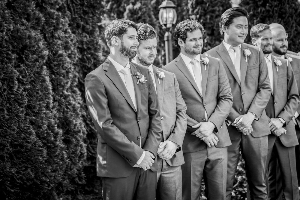 A group of groomsmen in formal suits share a candid laughing moment outdoors.