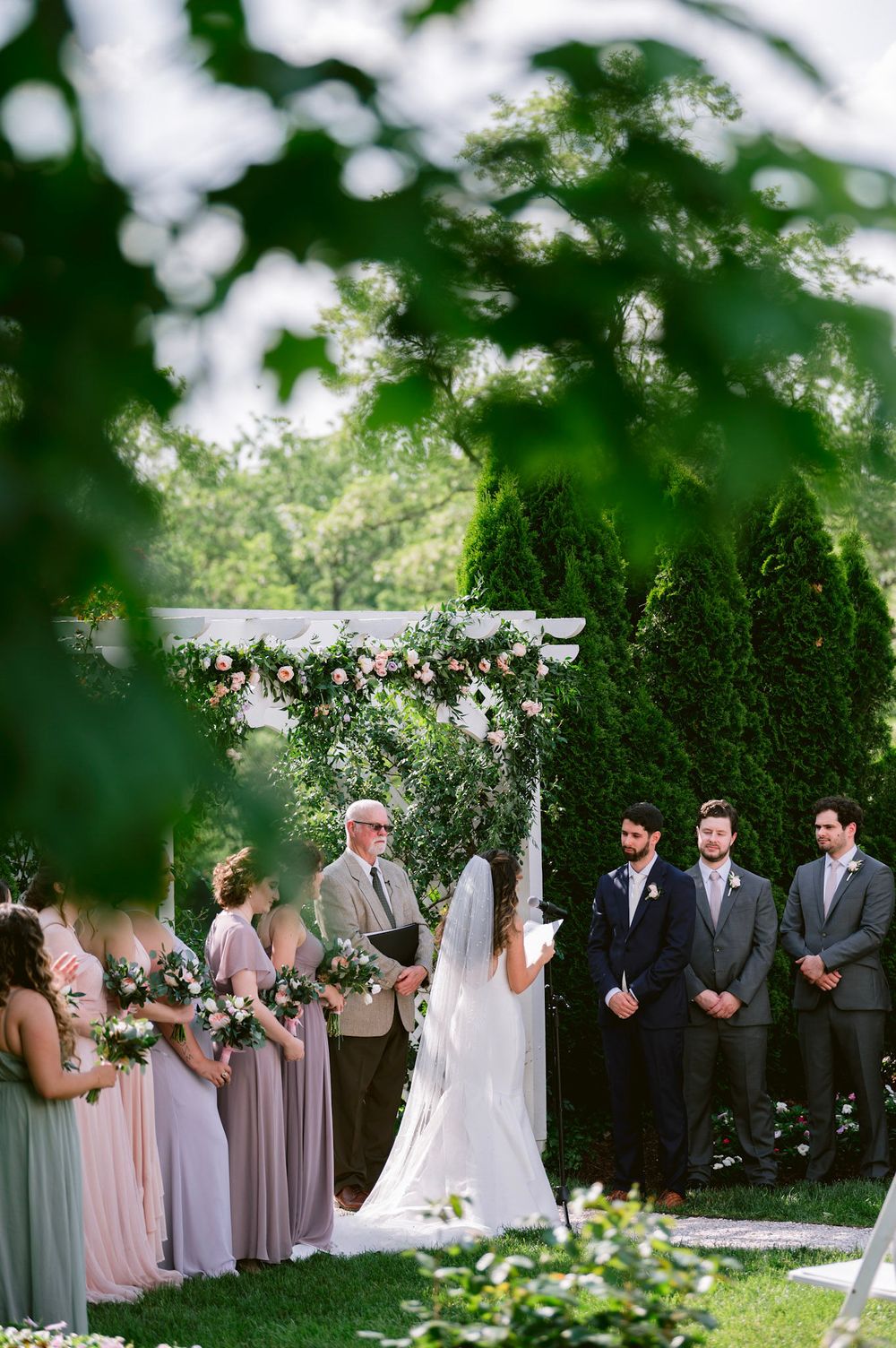 Wedding ceremony setup in a lush garden with white arch and floral decorations under green trees.