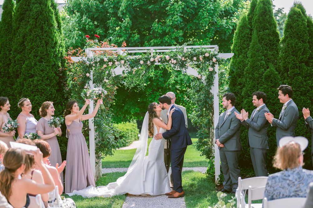 A romantic outdoor wedding ceremony takes place under a floral arch surrounded by manicured evergreen trees.