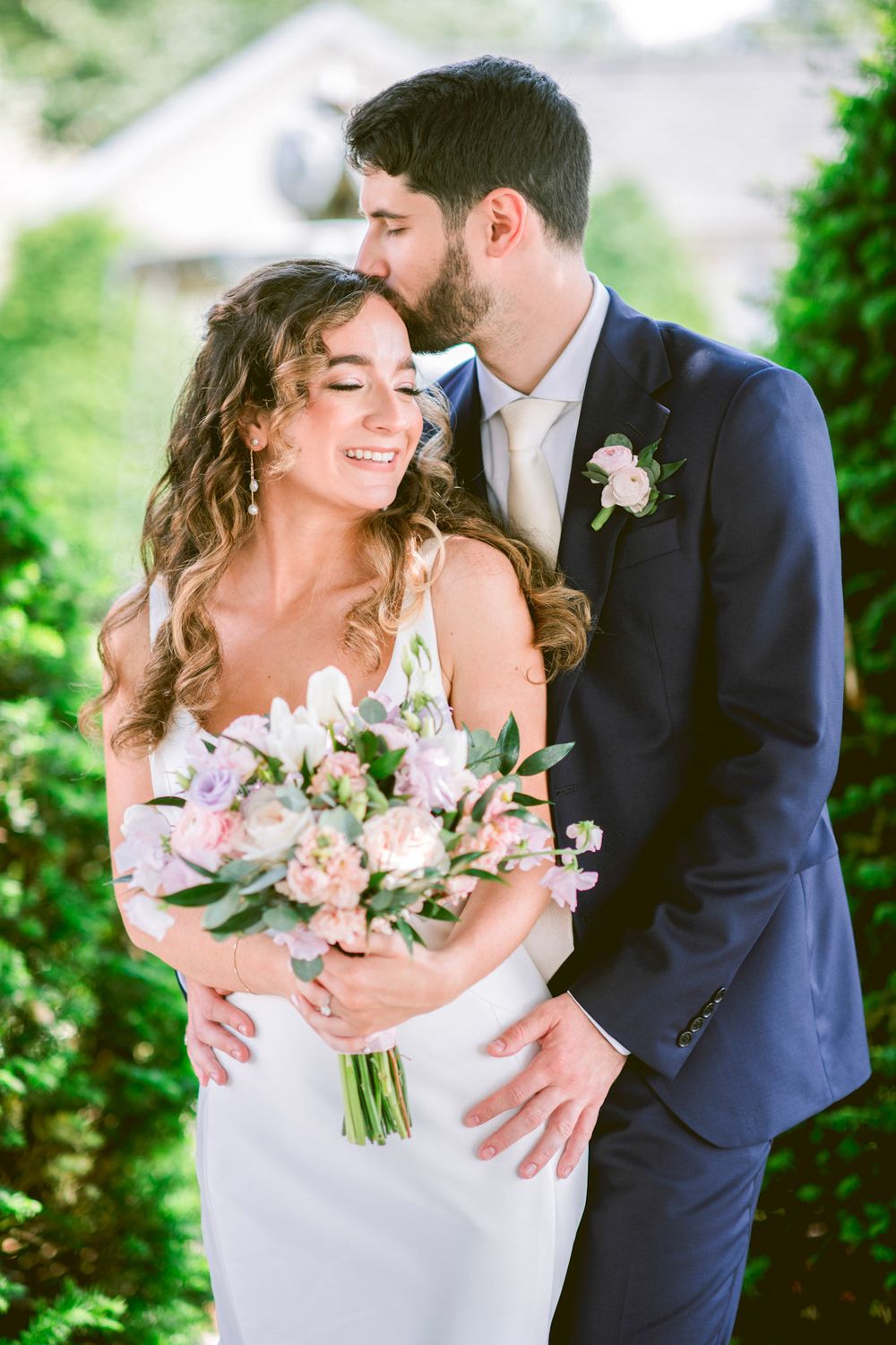 Romantic wedding couple embracing outdoors with pastel floral bouquet against lush greenery.