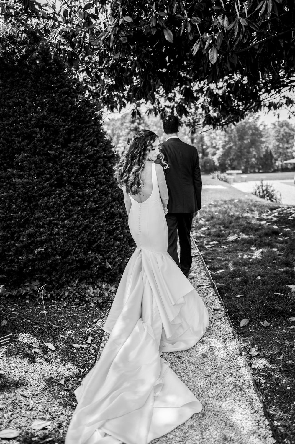 A black and white photo of a couple in wedding attire walking along a garden path under tree branches.